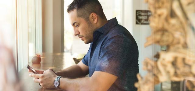 Man sitting at a long table in front of a window. He is looking at his phone.